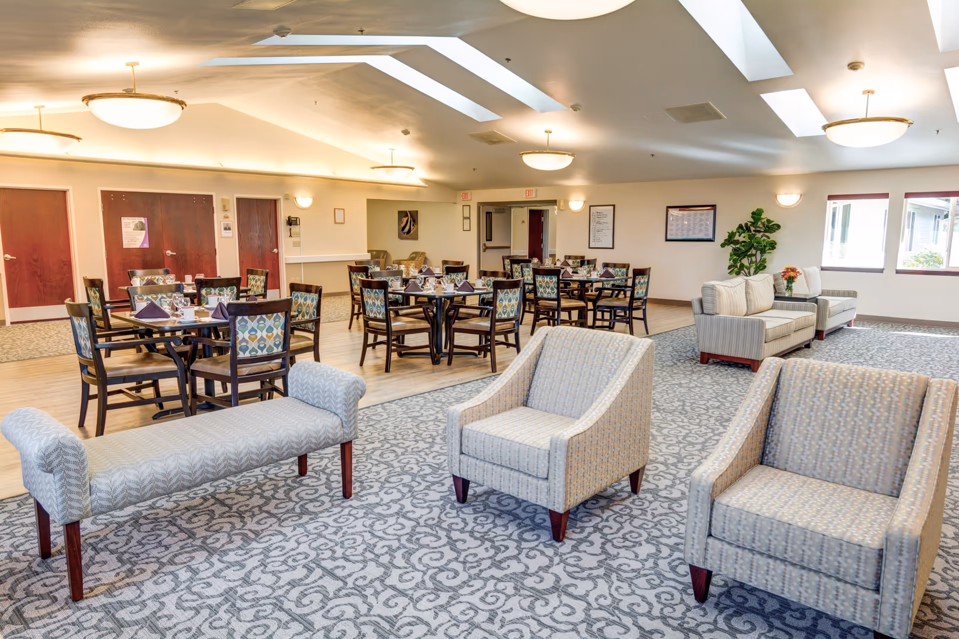 A spacious and well-lit common area in Monarch Gardens Memory Care featuring multiple dining tables with chairs arranged neatly, each table set with cups and napkins. The room also includes comfortable seating with armchairs and sofas on a patterned carpet, large windows allowing natural light, and ceiling lights with skylights.