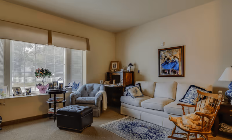 A cozy living room with a beige sofa adorned with blue and white pillows, a cushioned armchair, a wooden rocking chair with a doll, a small round table with framed photos and a flower vase, a window with blinds and a beige valance, a wooden cabinet, a table lamp, and a framed painting of dancers on the wall.
