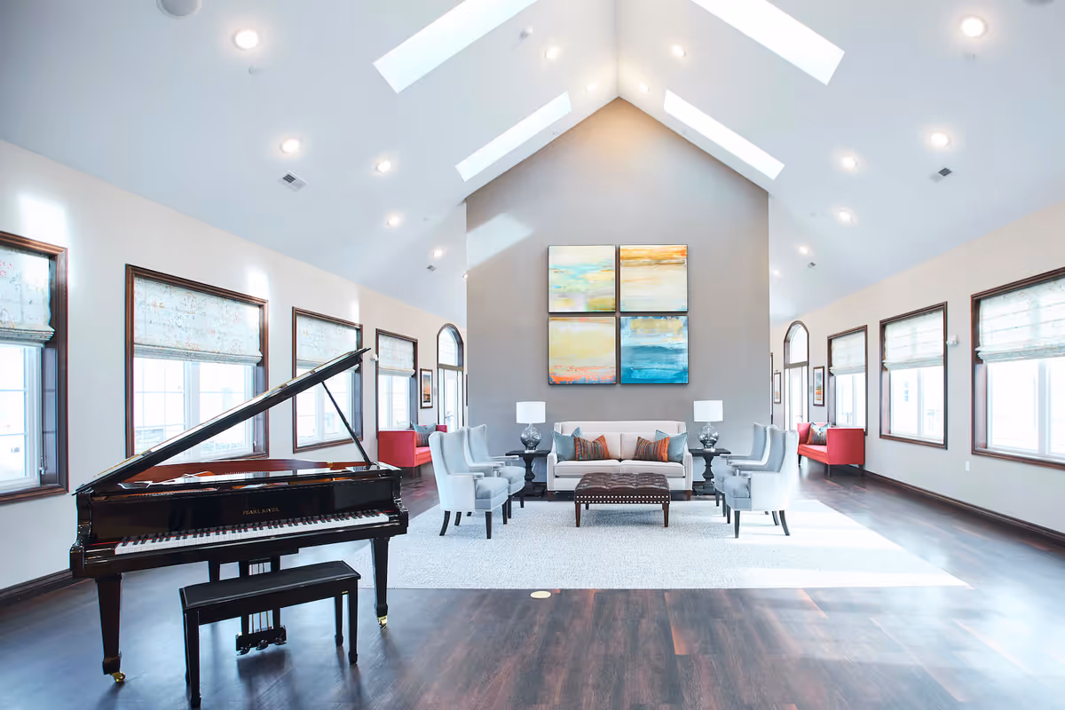 Bright and spacious living room with a high vaulted ceiling featuring skylights, a black grand piano on the left, a seating area with a beige sofa, four gray armchairs, two side tables with lamps, and colorful abstract paintings on the far wall. Large windows with patterned blinds line both sides of the room, and two red cushioned benches are placed near the windows.