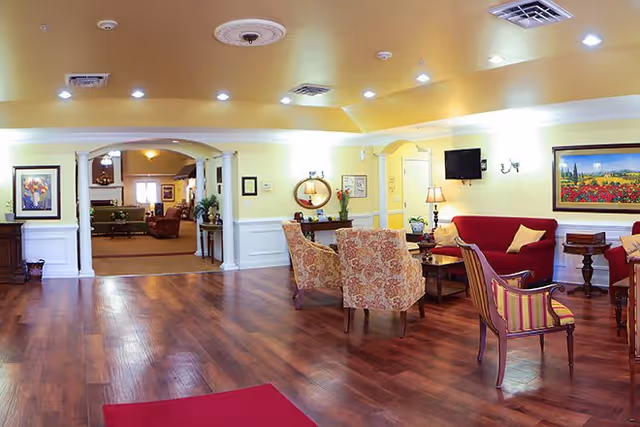 A spacious and well-lit living room area with wooden flooring, several upholstered chairs and sofas arranged around small tables, wall-mounted TV, framed artwork, and decorative lamps. The room has soft yellow walls and white trim, with an open archway leading to another seating area in the background.
