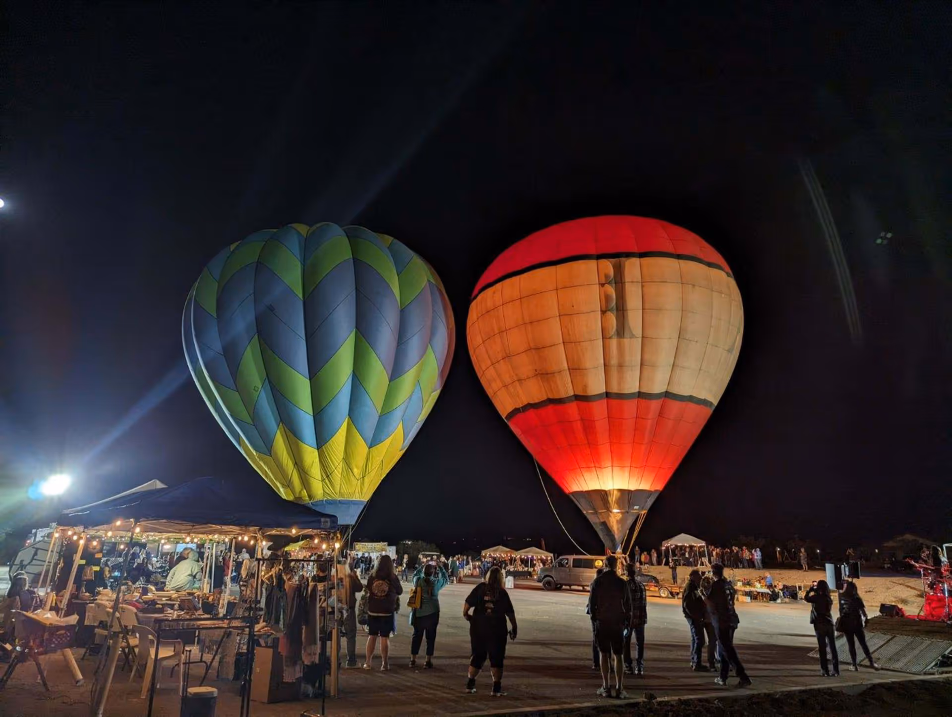 Nighttime scene of two illuminated hot air balloons on the ground with people standing nearby. There are tents and stalls with lights and merchandise on the left side, and a dark sky in the background.