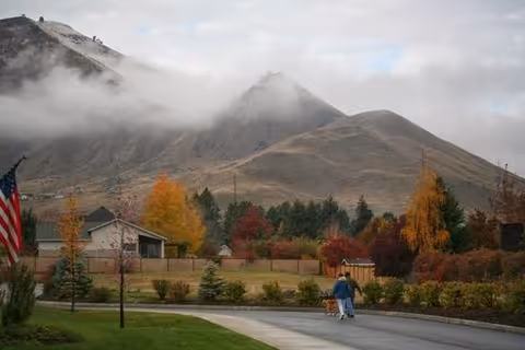 A person walking a dog on a paved path in a landscaped outdoor area with trees showing autumn colors. In the background, there are houses and large hills partially covered with low clouds or fog.