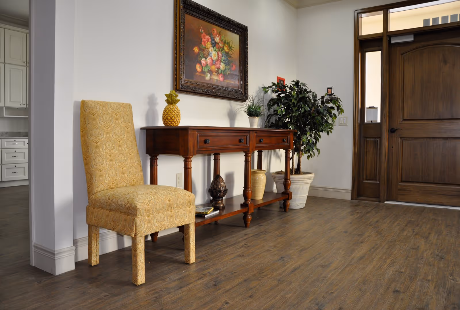 An entryway area with a wooden door, a wooden console table decorated with a pineapple ornament, a potted plant, and a framed floral painting on the wall. A yellow upholstered chair is positioned next to the console table, and the floor is covered with wood-style flooring.