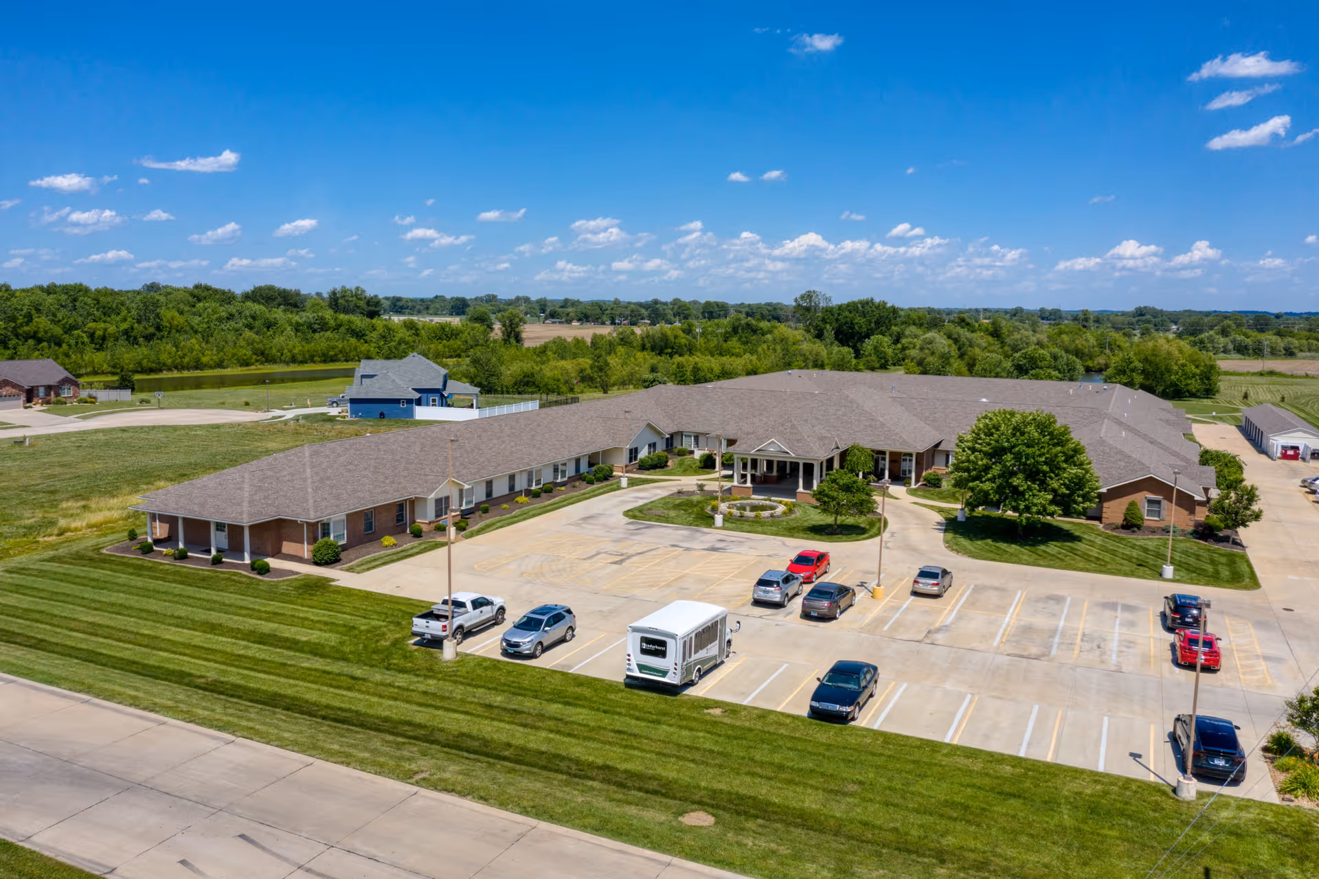 Aerial view of Cedarhurst Senior Living of Granite City, showing a large single-story building with a brown roof surrounded by green lawns and trees. There is a parking lot in front with several cars and a white shuttle bus parked. The sky is clear with a few clouds.