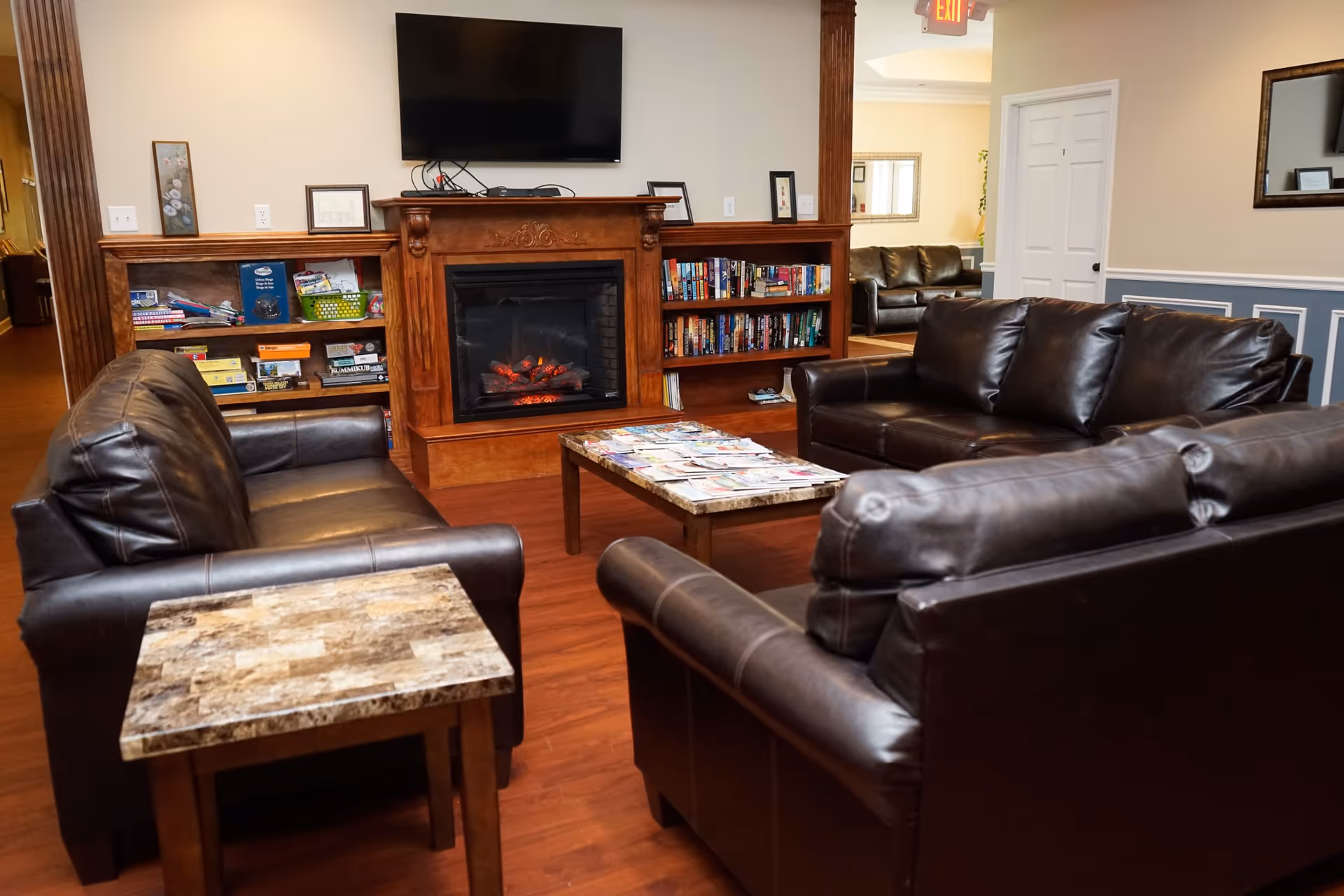 A cozy living room area with three dark brown leather sofas arranged around a rectangular coffee table covered with magazines. A wooden electric fireplace with a TV mounted above it is centered on the far wall, flanked by built-in bookshelves filled with books and games. The room has wood flooring and light-colored walls with decorative molding.