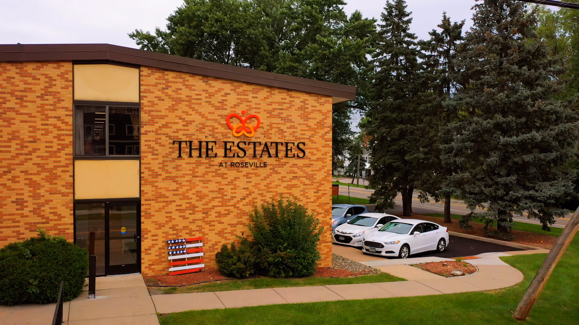 Exterior view of a brick building with the sign 'The Estates at Roseville' on the wall. There are bushes and trees around the building, a sidewalk leading to the entrance, and a small parking area with three white cars parked. The sky is partly cloudy.