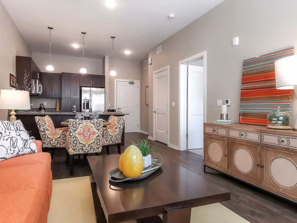 A modern senior living interior featuring a living room and kitchen area. The living room has an orange sofa with a white pillow decorated with black bunny illustrations, a dark wooden coffee table with decorative items, and a wooden sideboard with a colorful striped artwork and a lamp. The kitchen area has dark wood cabinets, stainless steel appliances, and three pendant lights hanging over a dining table with four patterned chairs. The walls are painted light beige, and the floor is dark wood.