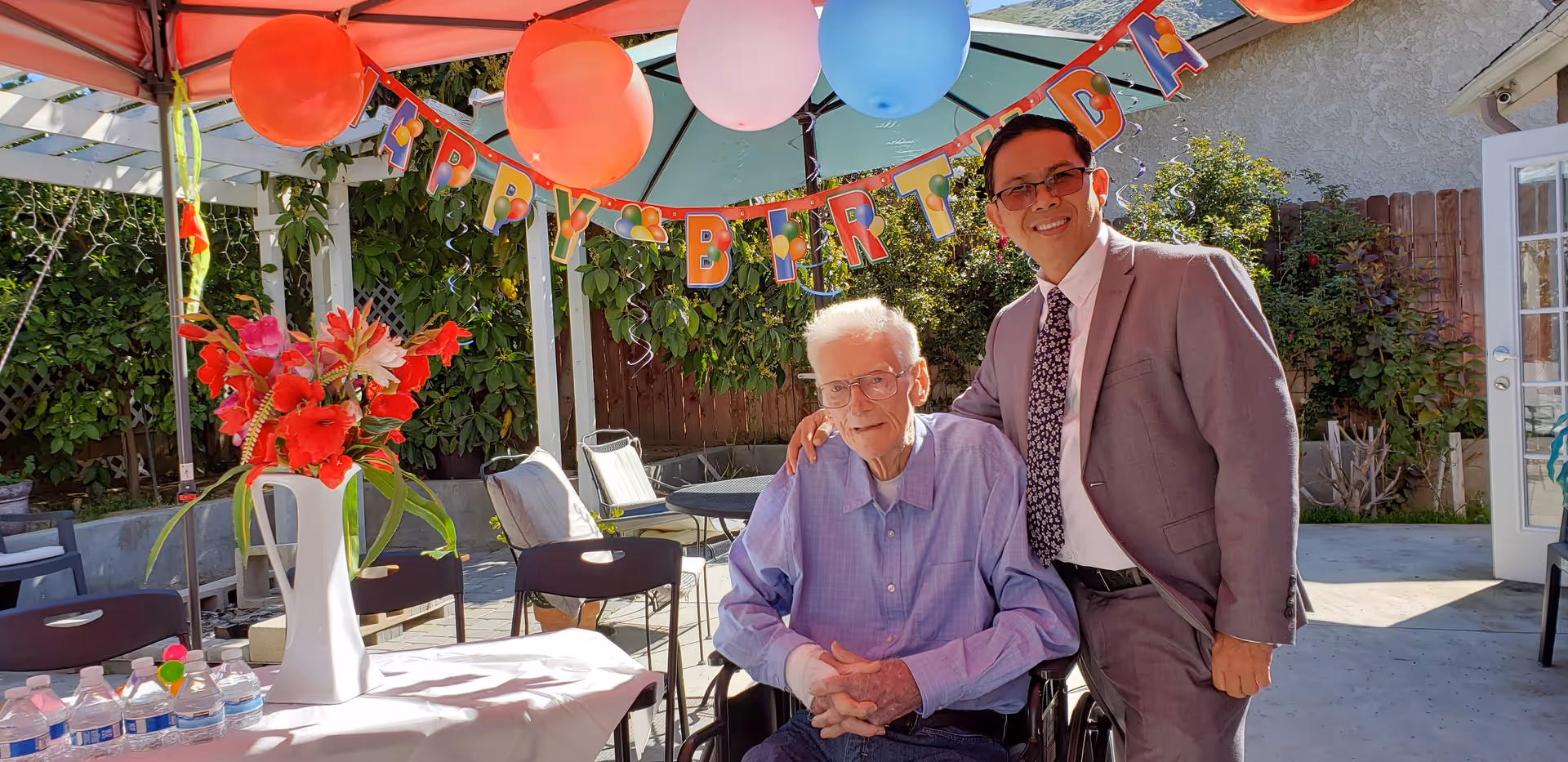 An elderly man in a wheelchair and a younger man in a suit smiling and posing together outdoors under a canopy decorated with colorful balloons and a 'Happy Birthday' banner. A table with a white tablecloth, a vase of red flowers, and several water bottles is visible in the foreground. Greenery and a wooden fence are in the background.