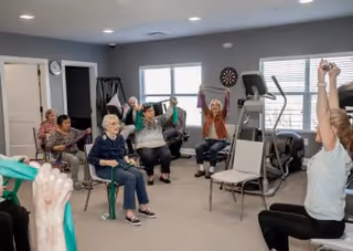 A group of elderly individuals seated in a room participating in a seated exercise session using green resistance bands, led by an instructor at the front. The room has exercise equipment, a dartboard on the wall, and large windows letting in natural light.
