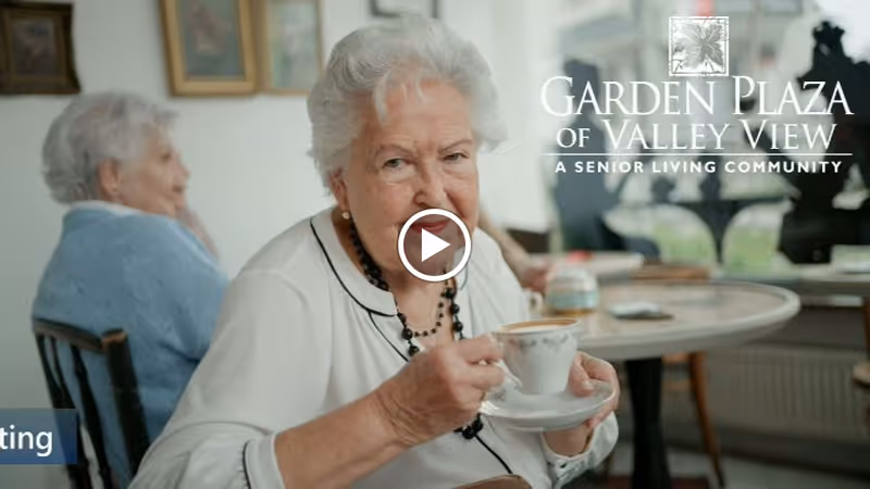 An elderly woman with white hair is sitting indoors at a table, holding a teacup and saucer, looking at the camera. Another elderly woman is seated in the background. The setting appears to be a cozy common area with framed pictures on the wall and a window letting in natural light.