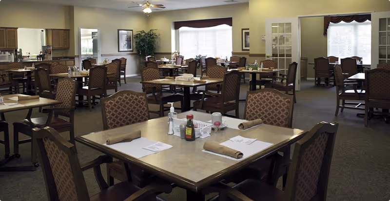 A spacious dining room in an assisted living facility with multiple tables and chairs arranged neatly. Each table is set with napkins, menus, and condiments. The room has large windows allowing natural light to brighten the space, and there are plants and framed pictures on the walls.