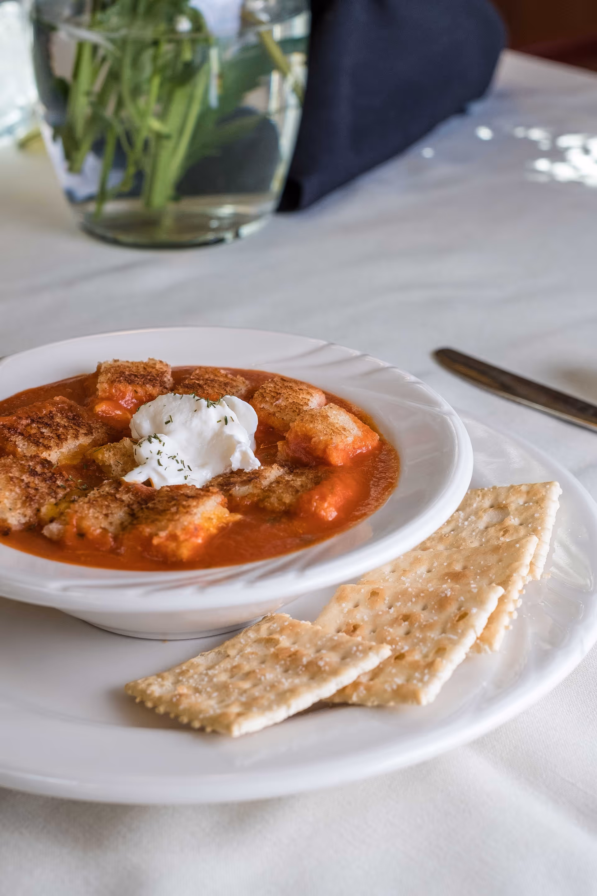 A bowl of tomato soup topped with croutons and a dollop of sour cream, served on a white plate with three saltine crackers on the side. A glass vase with green stems is visible in the background on a white tablecloth.