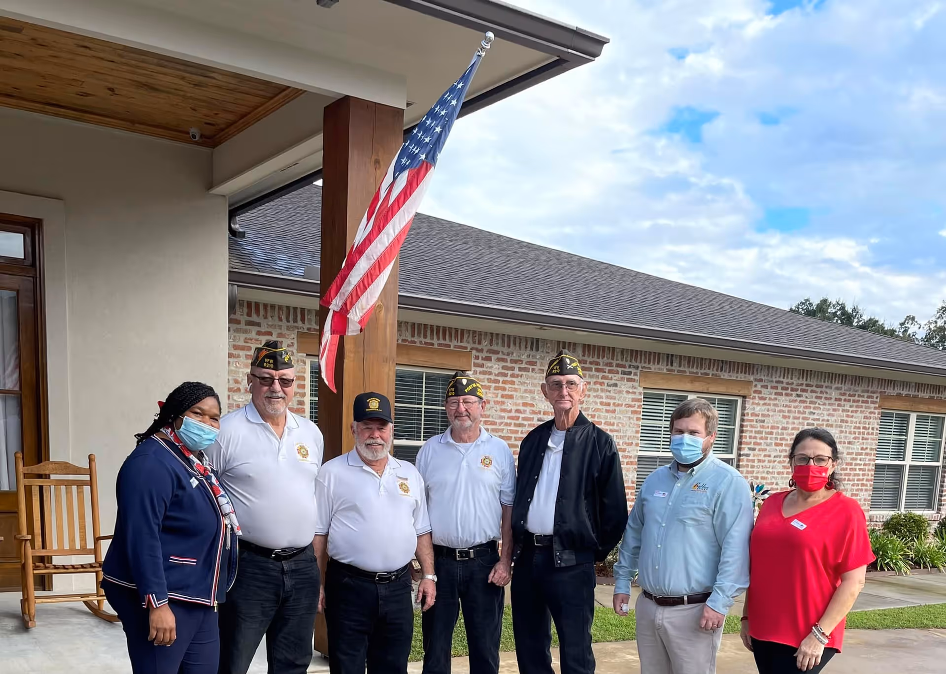 A group of seven people standing outside a brick building with an American flag mounted on a wooden post. Four men in white shirts and military hats stand in the middle, flanked by two people wearing masks and casual business attire on the right and one person wearing a mask and a navy blue outfit on the left. The setting appears to be the entrance of a senior living facility.