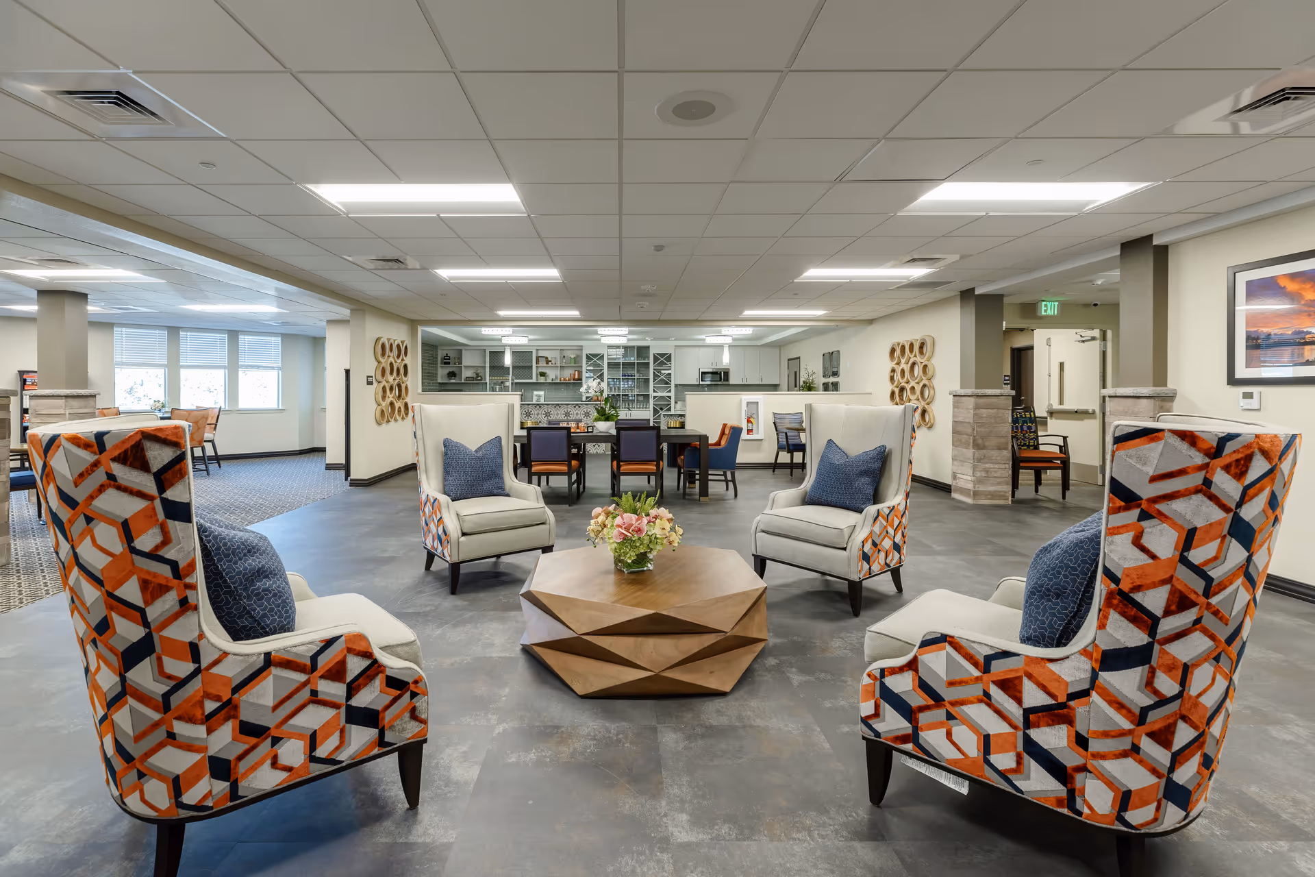 A spacious senior living common area with four patterned armchairs arranged around a geometric wooden coffee table with a flower arrangement. In the background, there is a dining area with tables and chairs, and a kitchen area with cabinets and appliances. The room has a tiled floor, a drop ceiling with fluorescent lights, and neutral-colored walls decorated with framed artwork and wall hangings.