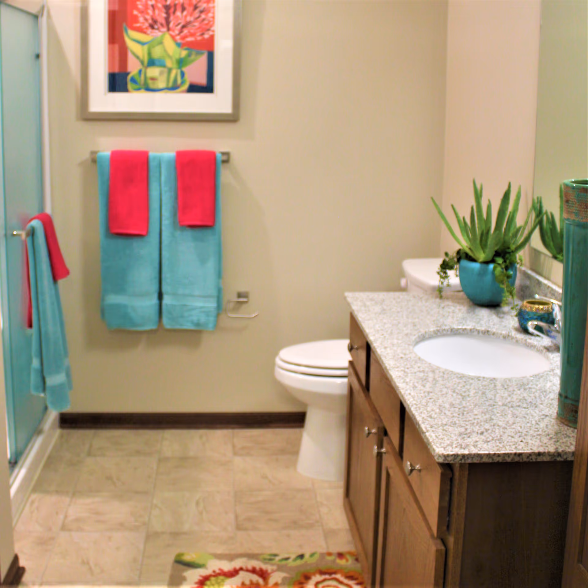 A clean bathroom with a granite countertop vanity featuring a sink, a potted green plant, and a decorative vase. There is a toilet next to the vanity, a glass shower door on the left, and two towel racks holding turquoise and red towels. A colorful floral painting hangs on the wall above the towels, and a patterned rug is on the tiled floor.