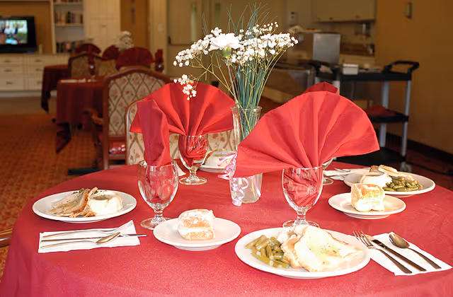 A dining table set with a red tablecloth, three plates of food including green beans, mashed potatoes, and rolls, three glasses with red folded napkins inside, and a vase with white flowers in the center. In the background, there are more tables and chairs in a dining area.