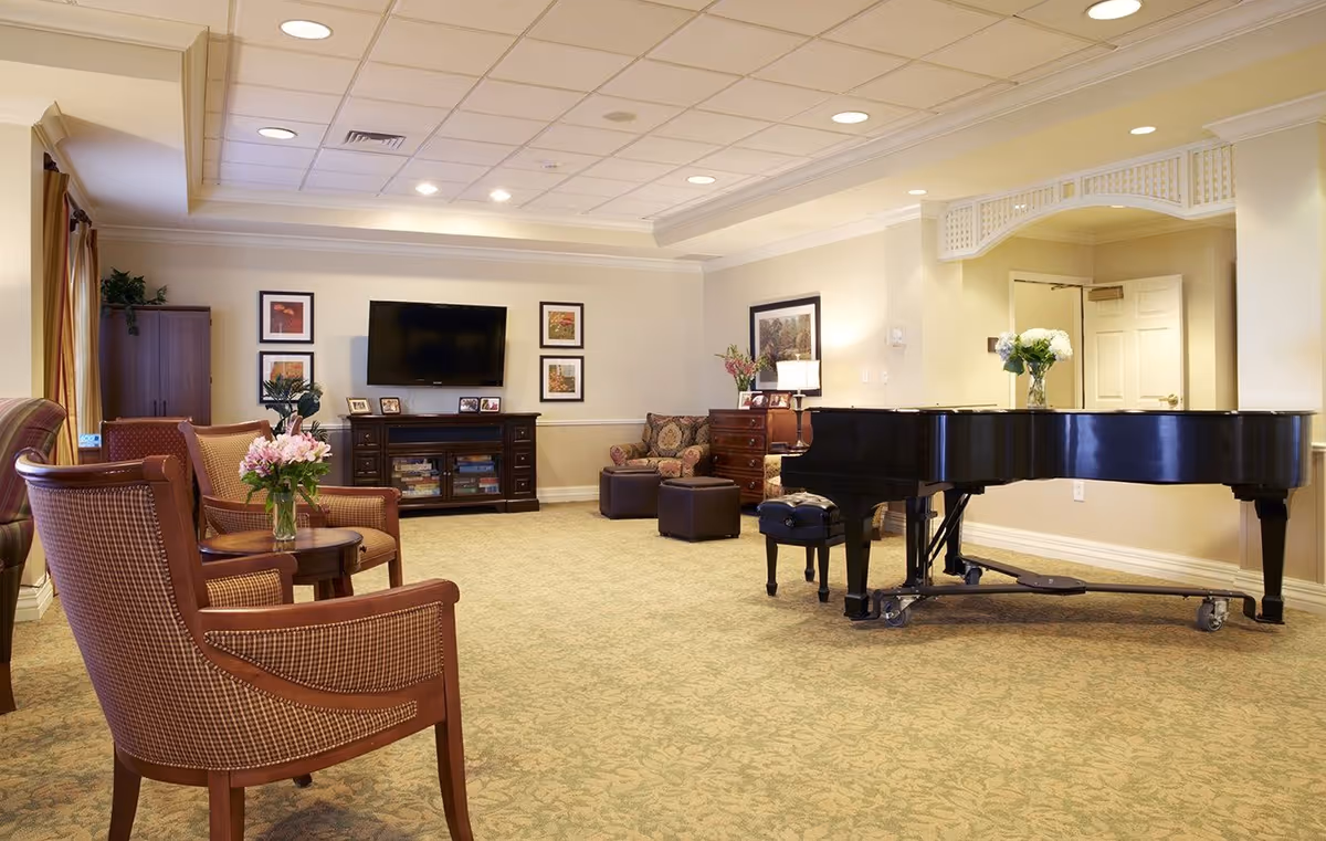 A spacious and well-lit senior living common area featuring a black grand piano with a vase of white flowers on top, several upholstered chairs with a small round table holding a vase of pink flowers, a flat-screen TV mounted on the wall above a wooden cabinet, framed artwork on the walls, and a cozy seating area with an armchair and ottomans.