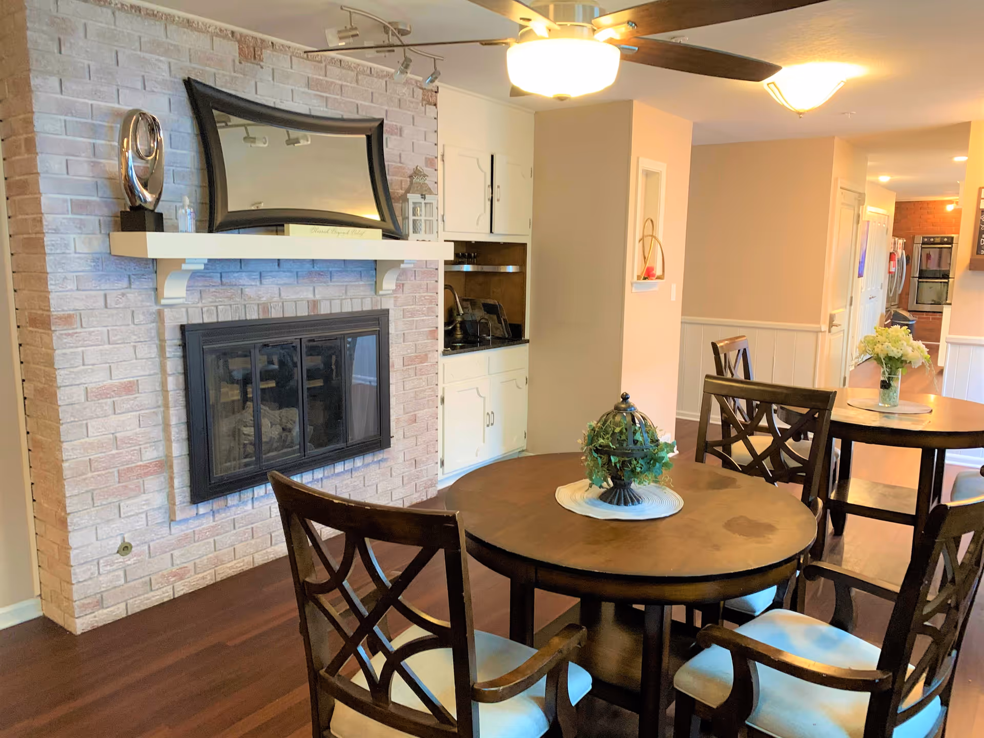 Interior view of a cozy common area with a round wooden table and four chairs with cushioned seats. A decorative centerpiece is on the table. There is a brick fireplace with a black frame and a white mantel holding a curved mirror and a silver decorative object. The room has wooden flooring, beige walls, a ceiling fan with a light, and a small built-in cabinet area with a sink. In the background, another round table with chairs and a vase of flowers is visible, along with a hallway leading to a kitchen area.