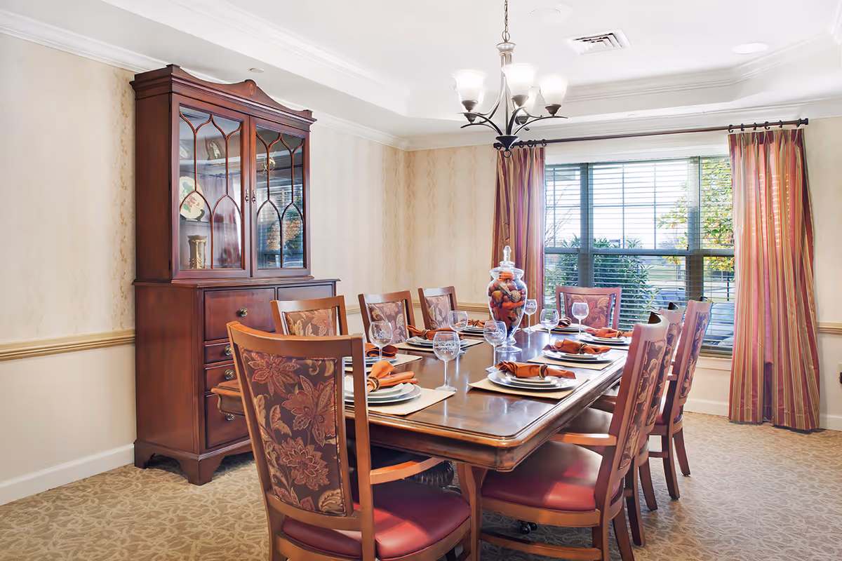 A formal dining room with a wooden table set with place settings, upholstered chairs, a china cabinet, and a window with curtains.