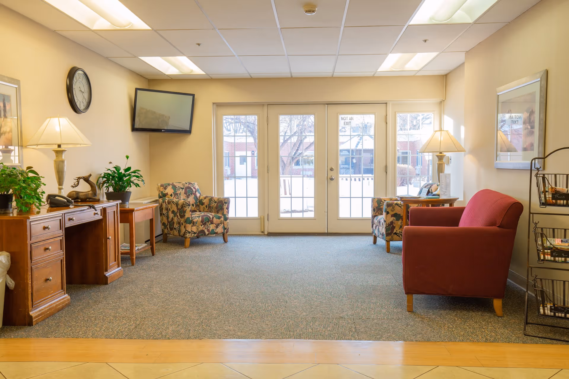 A bright and cozy common area with a red sofa, two patterned armchairs, a wooden sideboard with plants and a lamp, a wall clock, a wall-mounted TV, and double glass doors leading outside. The room has beige walls, carpeted floor, and framed artwork on the walls.
