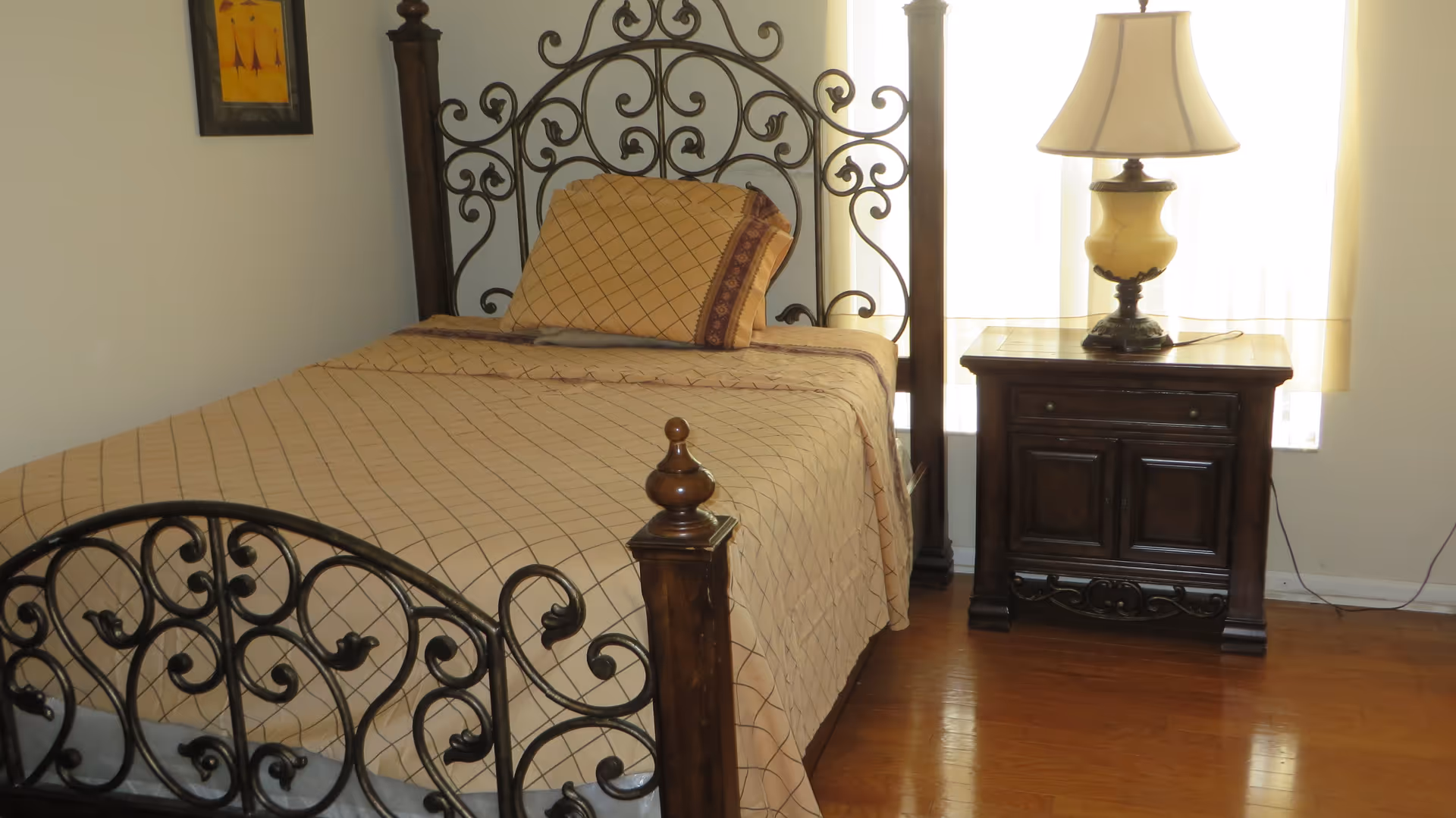 A bedroom with a single bed featuring an ornate wrought iron headboard and footboard. The bed is made with a beige bedspread and matching pillow with a checkered pattern. Next to the bed is a dark wooden nightstand with a decorative lamp on top. A window with light curtains is behind the nightstand, and a framed picture hangs on the wall above the bed.