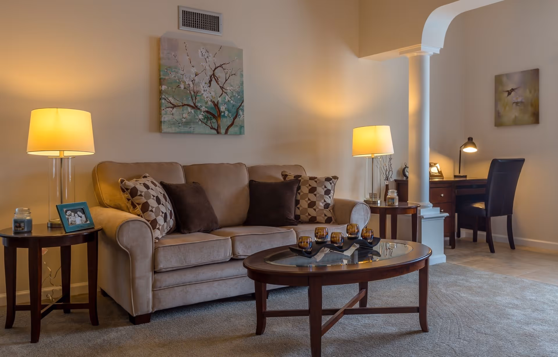 A cozy living room with a beige sofa adorned with patterned and solid dark pillows, two side tables each with a lamp, a glass-top coffee table with decorative candle holders, a framed photo, and artwork on the walls. In the background, there is a small desk with a chair and a desk lamp, separated by a white column.