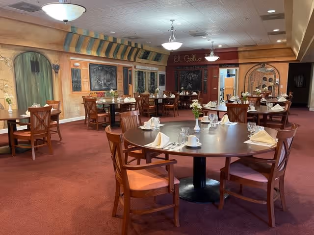 Spacious dining room featuring round wooden tables set for meals, wooden chairs, carpeted floor, and mural-decorated walls.
