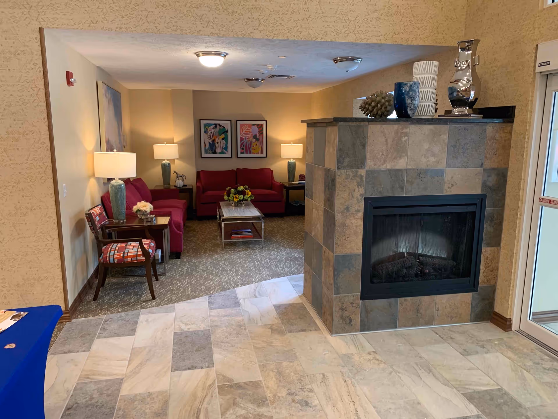 A cozy indoor lounge area with a tiled fireplace in the foreground. Behind the fireplace, there are two red sofas, a coffee table with a flower arrangement, two side tables with lamps, and two framed abstract paintings on the wall. A patterned armchair and a small table with a lamp are positioned to the left. The floor near the fireplace is tiled, transitioning to carpet in the seating area. The walls are painted beige, and there is a glass door to the right.