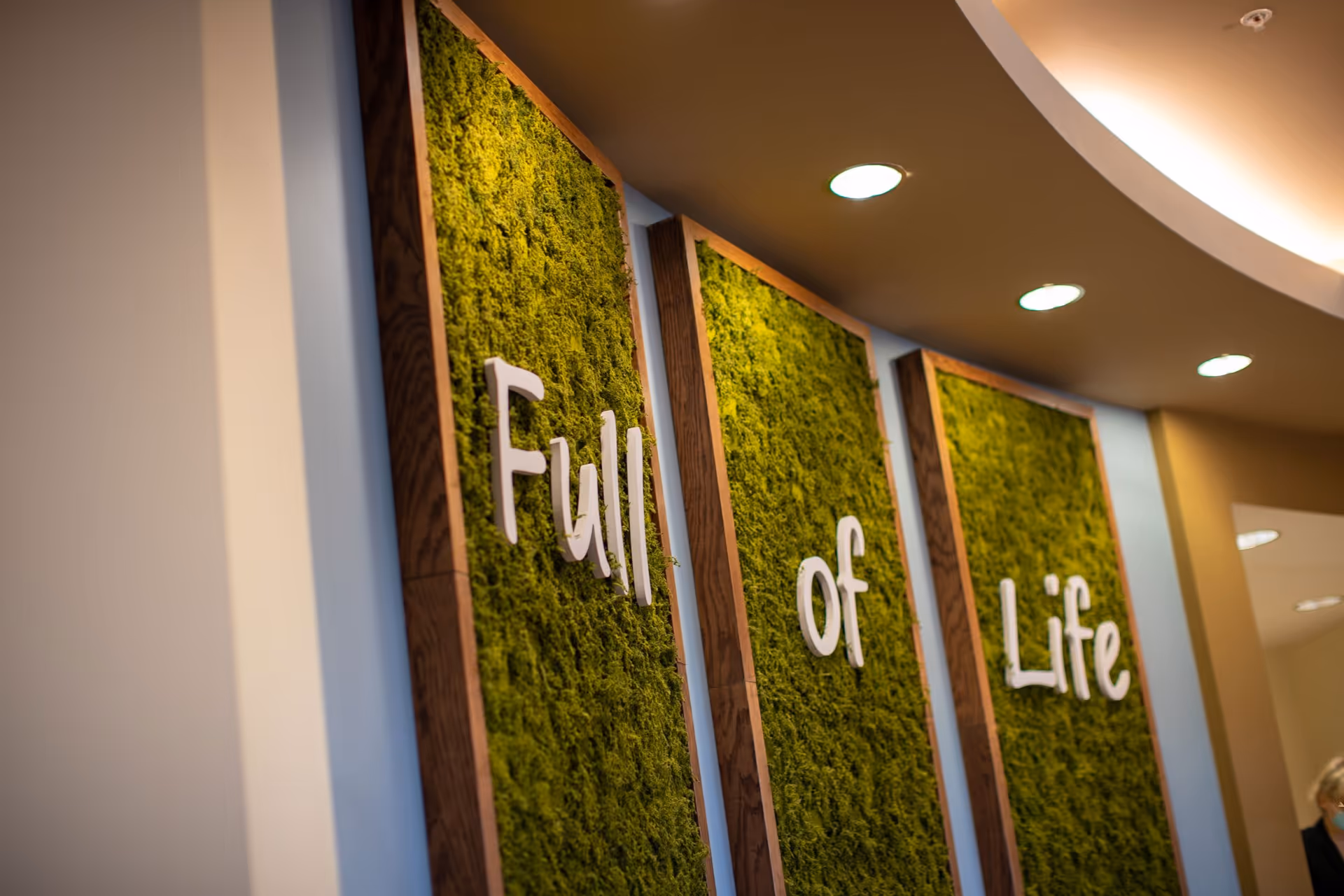 Three vertical panels covered with green moss mounted on a wall, each framed in wood, with white letters spelling out the phrase 'Full of Life'. The ceiling above has recessed lighting.