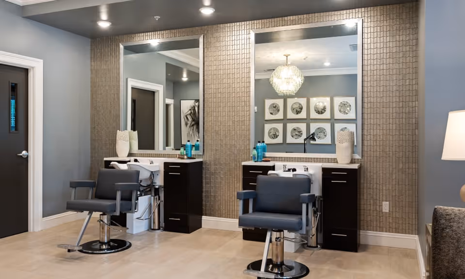 Interior view of a salon area with two black salon chairs in front of two large mirrors mounted on a textured wall. Each station has a white sink and dark cabinetry with hair care products on the counter. The room has light-colored flooring, a gray door, and a lamp on the right side.
