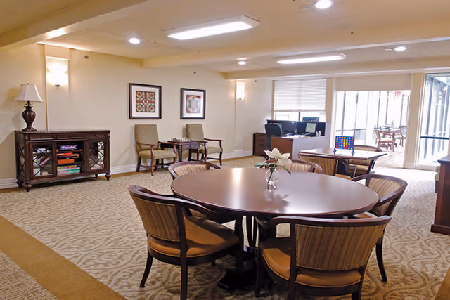 A well-lit common area in a senior living facility featuring a round wooden table with six chairs, a small vase with flowers on the table, two armchairs with a small table between them against the wall, framed artwork on the wall, a wooden cabinet with books and a lamp, and a desk with a computer near large windows and a glass door leading to an outdoor patio.