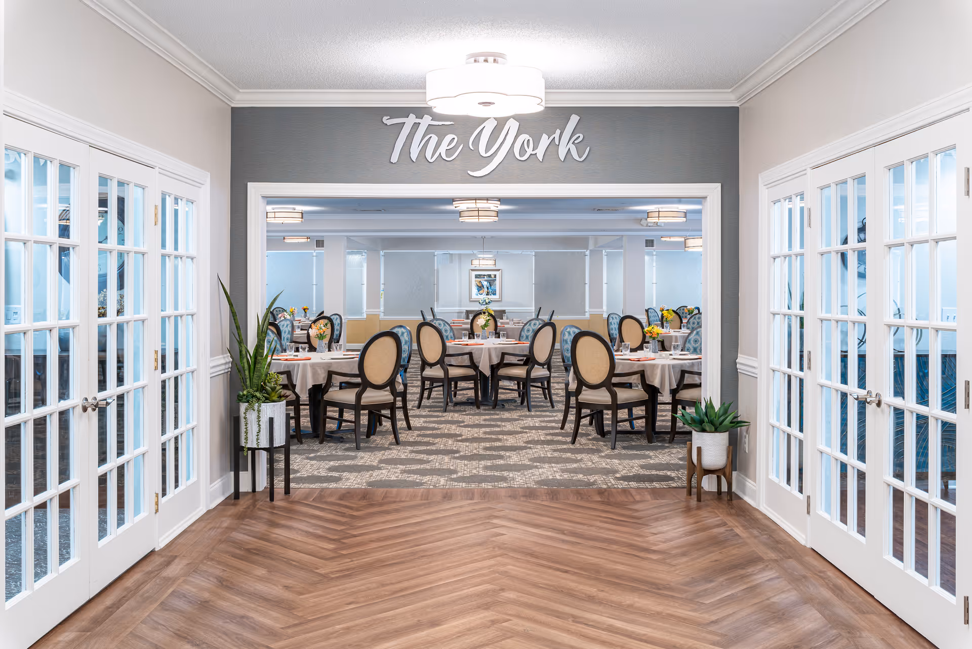 View through a hallway with wooden herringbone flooring leading to a dining room named 'The York'. The dining room has round tables covered with white tablecloths, each surrounded by elegant chairs with beige upholstery and dark wooden frames. The room is well-lit with ceiling lights and decorated with plants on either side of the hallway entrance.