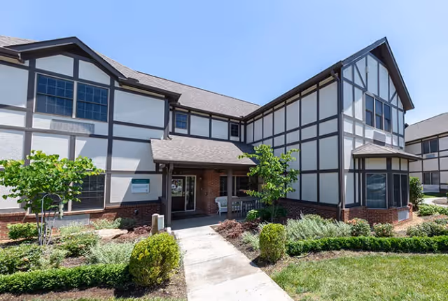 Exterior view of a two-story senior living facility building with Tudor-style architecture, featuring white walls with dark wooden beams, a shingled roof, and a landscaped entrance pathway surrounded by bushes and small trees under a clear blue sky.