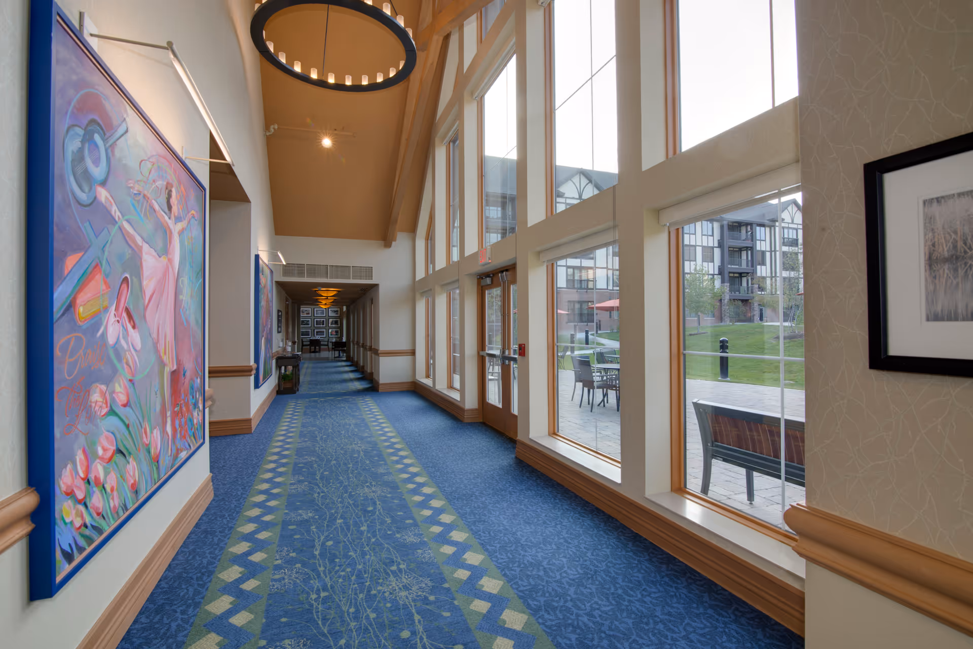A long, carpeted hallway with large windows on the right side showing an outdoor patio area with tables and chairs. The left wall features colorful artwork, and the ceiling is high with a circular light fixture. The hallway leads to a seating area at the far end.