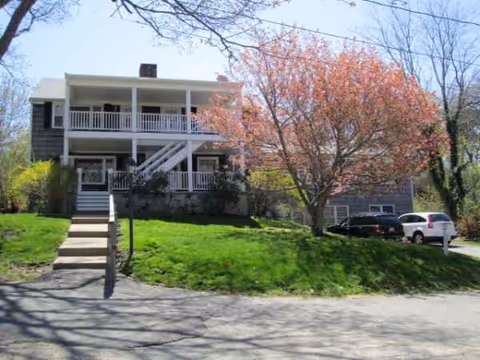 Two-story senior living building with white porches, stairs, green lawn, and parked cars.