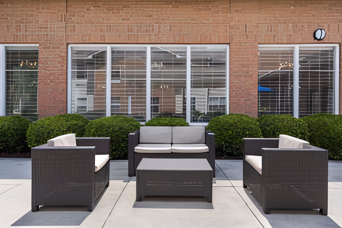 Outdoor patio area with modern dark wicker furniture including two armchairs, a loveseat, and a coffee table, set on a concrete surface in front of a brick building with large windows and green bushes.