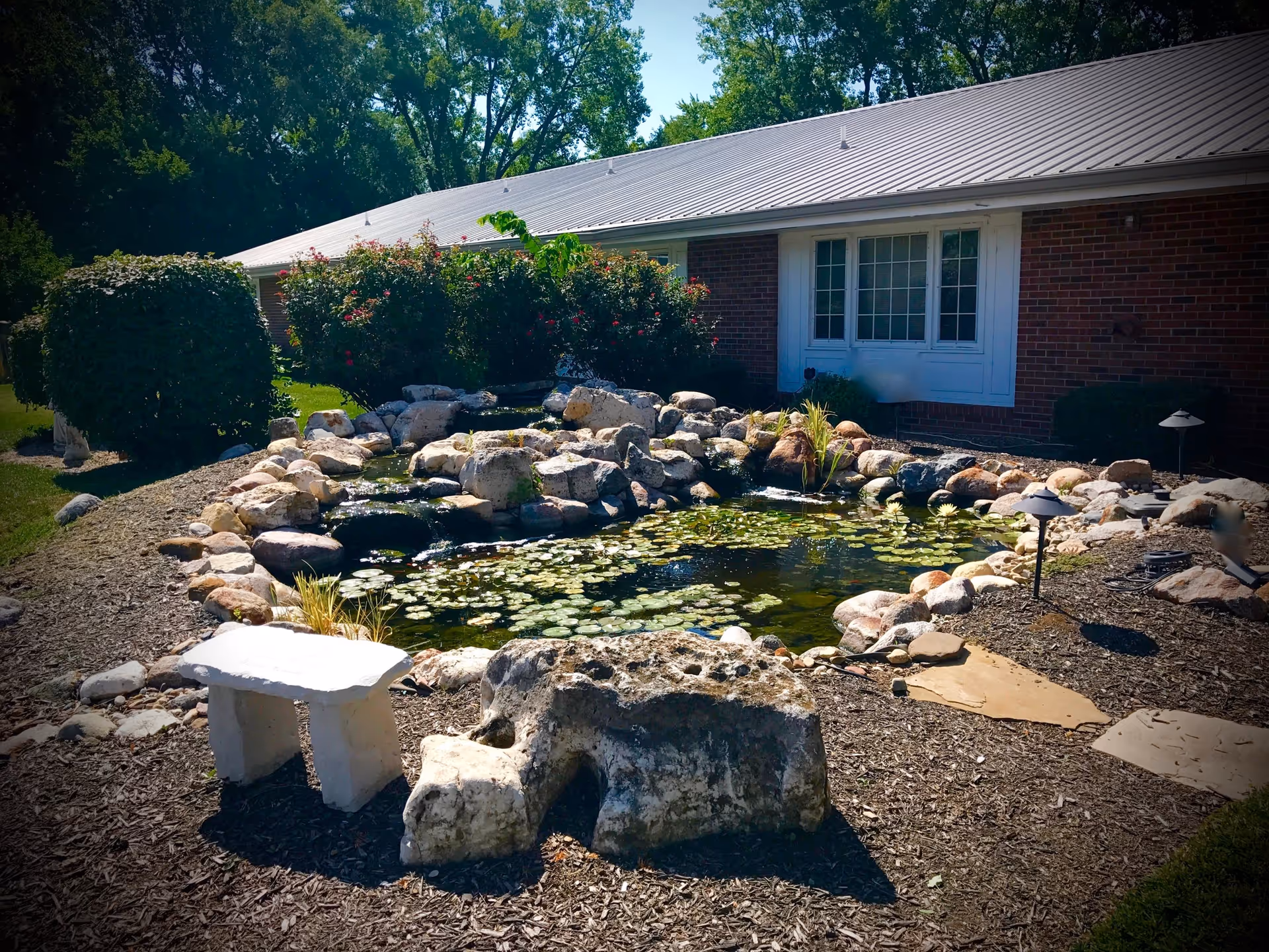 A landscaped outdoor area featuring a small pond with lily pads surrounded by rocks. There is a white stone bench and a large rock in the foreground. Behind the pond, there are bushes and a brick building with white-framed windows. Trees are visible in the background under a clear sky.