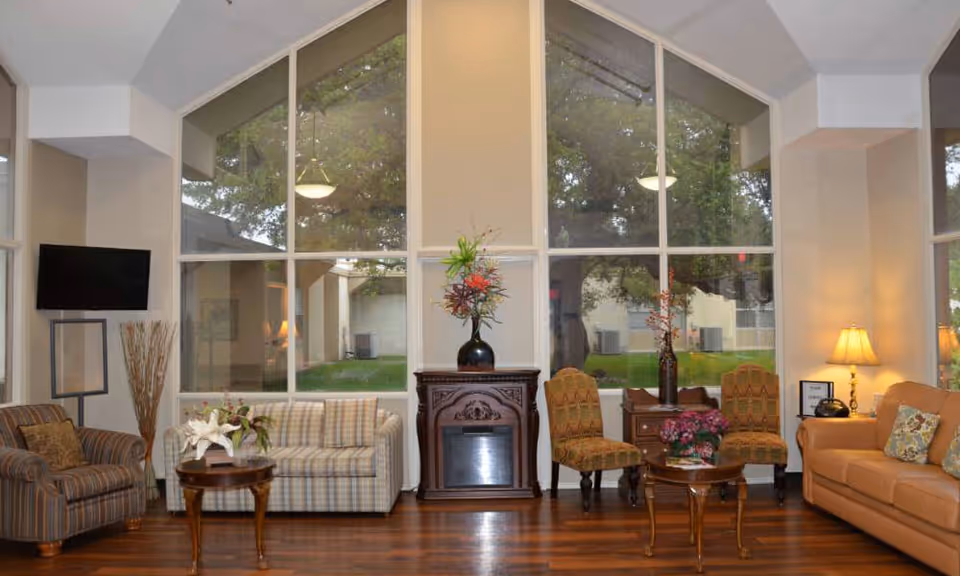 A cozy living room area with large floor-to-ceiling windows showing trees outside. The room features a striped armchair, a plaid sofa, two patterned chairs, a tan leather sofa, wooden tables with floral arrangements, a decorative fireplace, a floor lamp, and a wall-mounted TV.