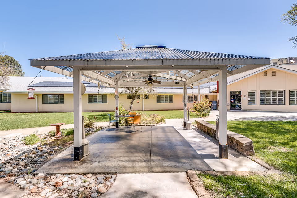 Outdoor covered patio area with a metal roof and ceiling fan, featuring a small table with chairs underneath. The patio is surrounded by a well-maintained lawn, landscaping with rocks, and a beige building with multiple windows in the background under a clear blue sky.