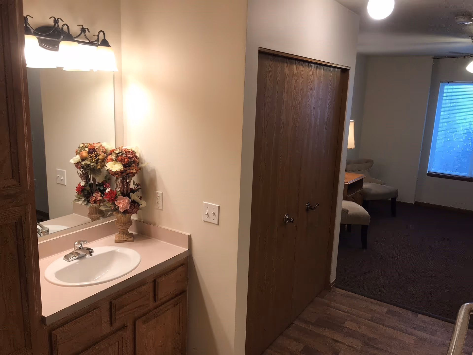 A vanity with a sink, mirror and floral arrangement beside closed wooden closet doors opening into a small living area with chairs and a window.