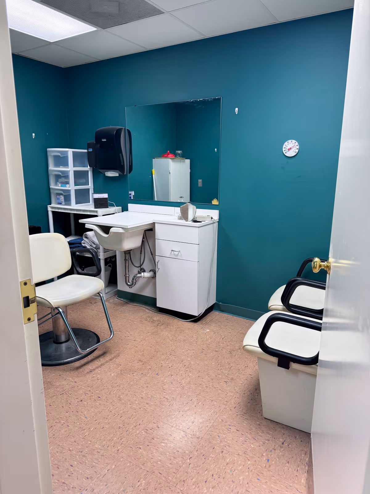 Small room with teal walls featuring a white sink with cabinet, a large mirror above the sink, a white chair with metal legs, two white chairs with black armrests, a plastic drawer organizer, a wall-mounted black paper towel dispenser, and a round wall clock.