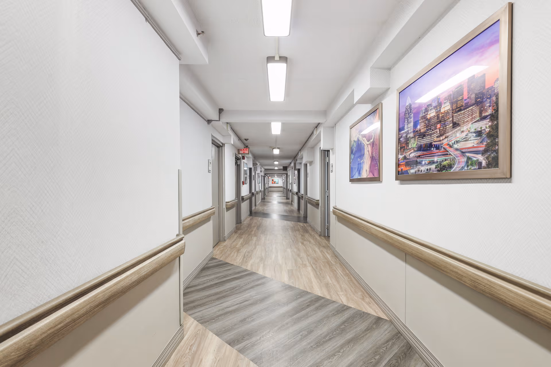 A long, well-lit hallway in a healthcare facility with light-colored walls and wood-patterned flooring. The hallway has handrails on both sides and framed artwork hanging on the right wall. Several doors line the corridor, and ceiling lights illuminate the space evenly.