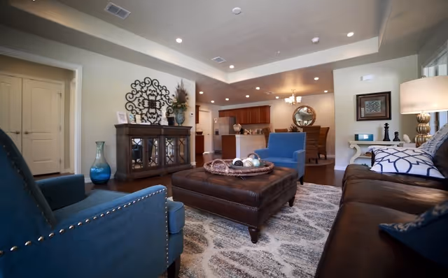 A cozy living room with blue armchairs, a brown leather sofa, and a large brown leather ottoman in the center. The room features a patterned area rug, a wooden cabinet with decorative items, and a lamp on a side table. In the background, there is an open kitchen and dining area with wooden cabinets and a round mirror on the wall.