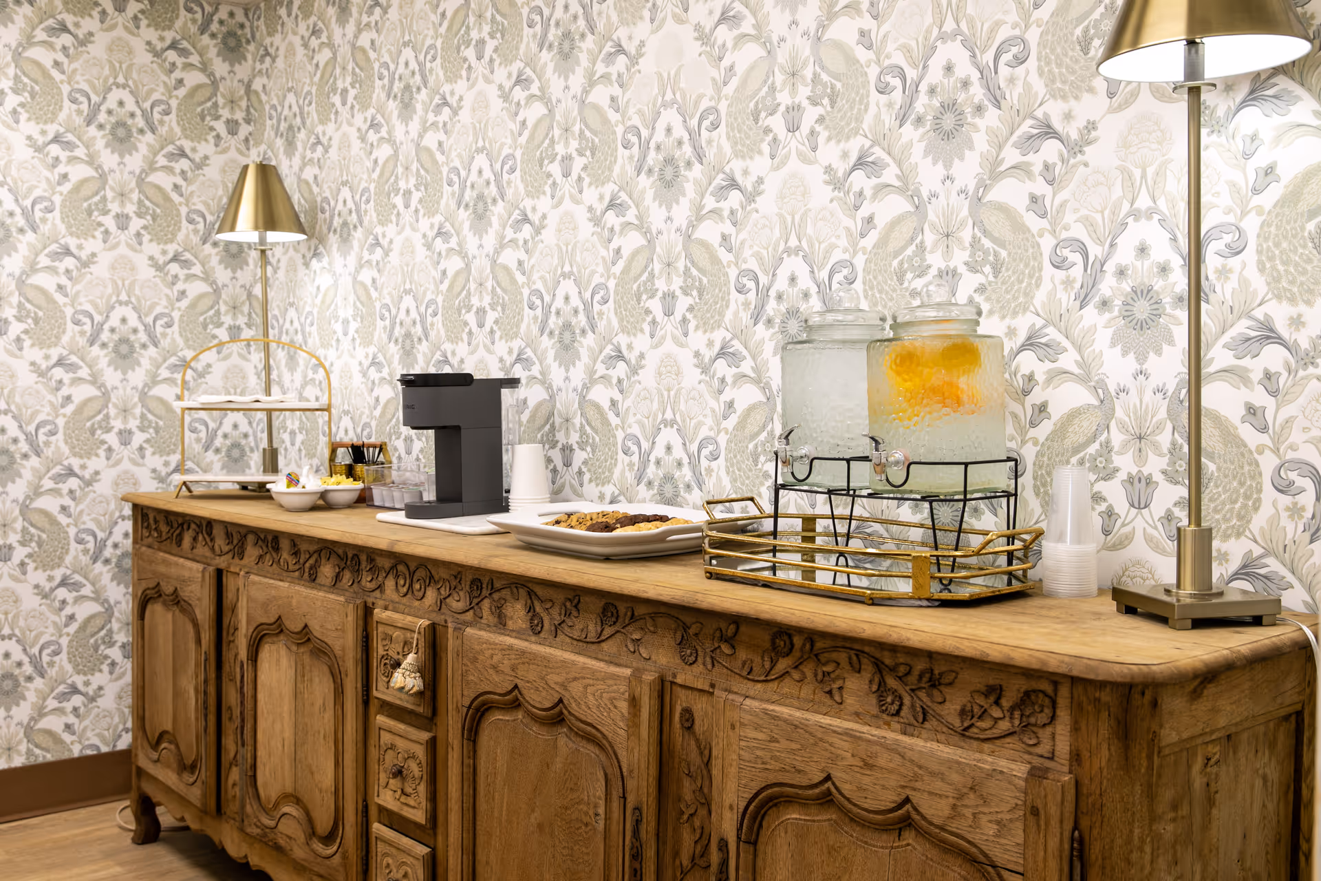 A wooden sideboard with intricate carvings against a wall with floral and peacock patterned wallpaper. On the sideboard are two glass beverage dispensers, a coffee maker, a plate of cookies, small bowls with snacks, and two brass table lamps with white shades.