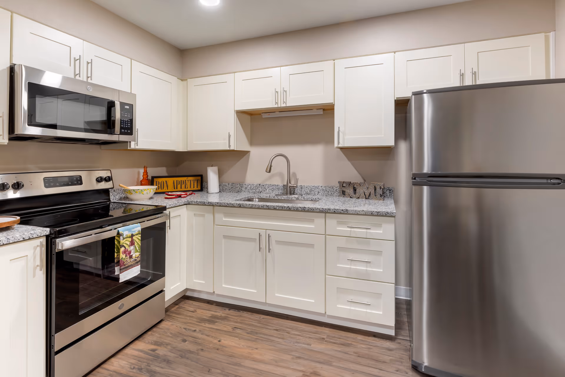 Modern kitchen with white cabinets, granite countertops, stainless steel appliances including a microwave, stove, and refrigerator. The kitchen has a wooden floor and decorative items such as a bowl, a 'Bon Appetit' sign, and a 'HOME' sign on the countertop.