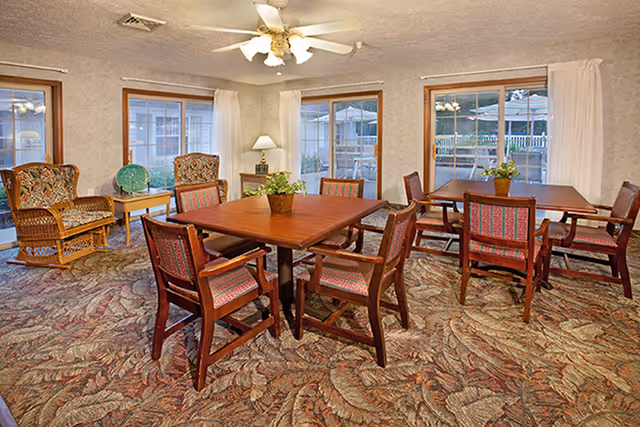 A cozy common area with two wooden tables surrounded by cushioned chairs with patterned upholstery. The room has large windows with white curtains, a ceiling fan with lights, a side table with a lamp and decorative globe, and two wicker chairs with cushions. The carpet has a leafy pattern in warm tones.