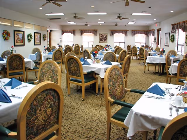 A spacious dining room with multiple tables covered in white tablecloths, each set with blue folded napkins, silverware, and small floral centerpieces. The chairs have wooden frames with floral upholstered backs and green seats. The room is well-lit with ceiling lights and natural light from windows with valances. The walls are decorated with framed pictures and wreaths.