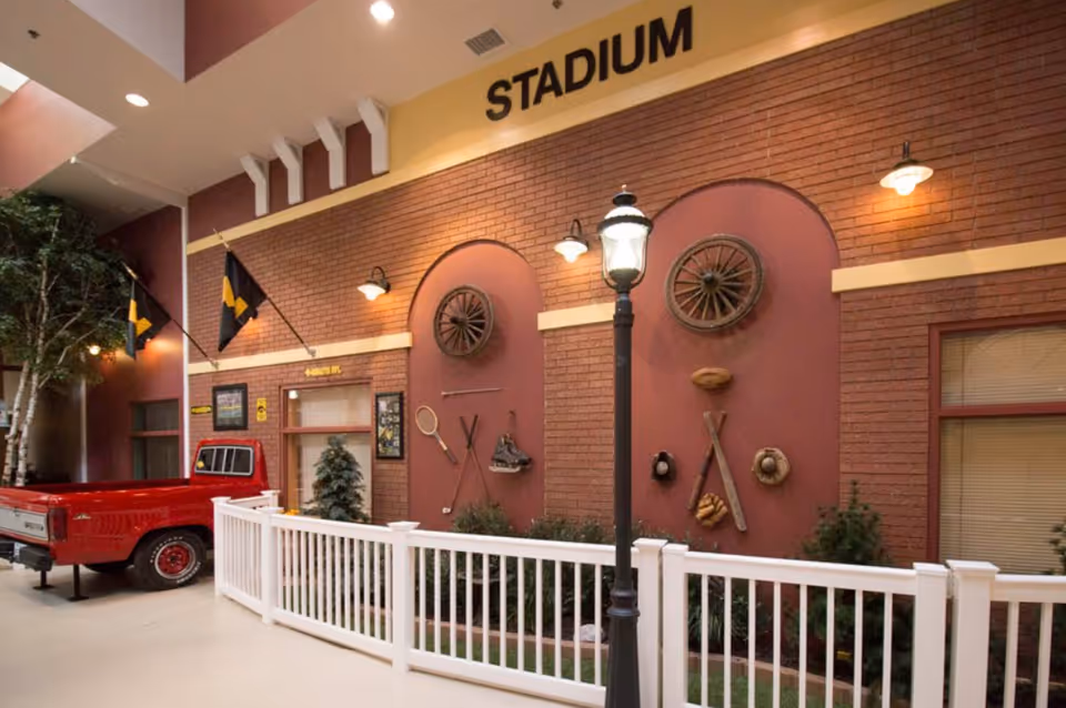 Indoor mock stadium-style facade with a brick wall decorated with sports memorabilia, a streetlamp, white fence, and a red pickup truck.