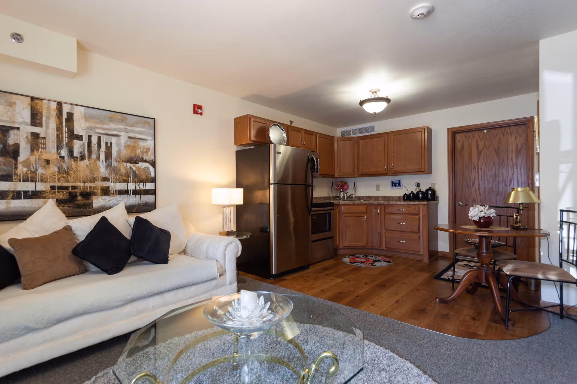 Open living area with a white sofa and glass coffee table next to a kitchenette with wooden cabinets and a stainless-steel refrigerator.