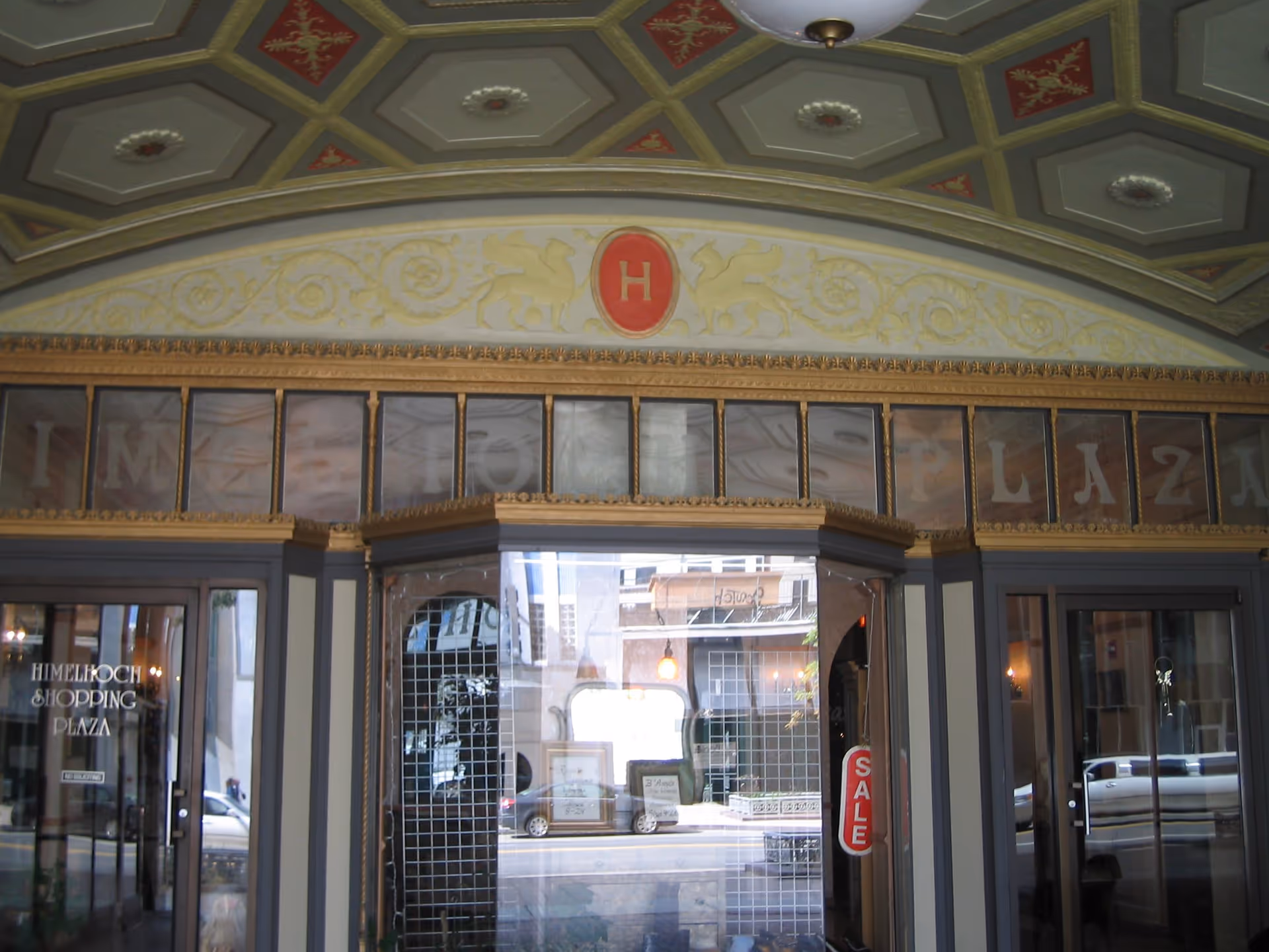 Entrance to Himelhoch Shopping Plaza with ornate architectural details including a decorative ceiling and a red emblem with the letter H above the doorway. Glass doors and windows reflect the street outside, and a red SALE sign is visible on one of the doors.